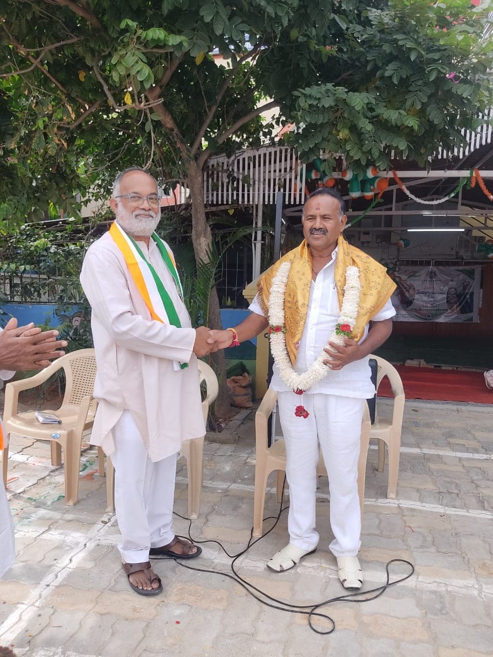 Two men shaking hands outdoors under a tree, one wearing a garland of flowers.