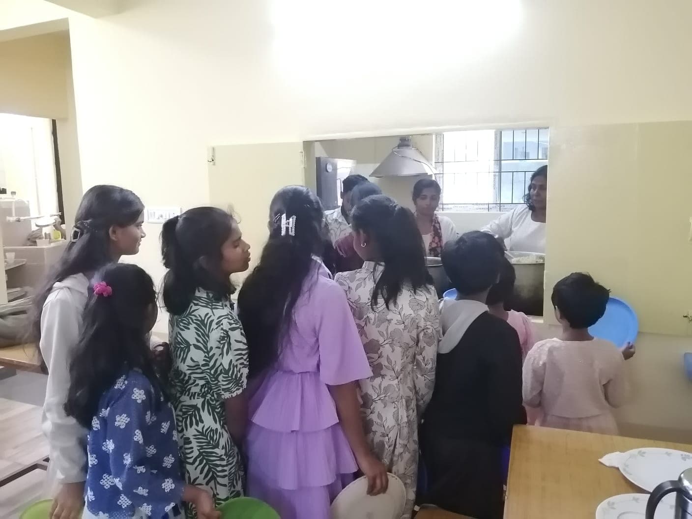 Children queuing with plates at a serving hatch while a cook serves rice.