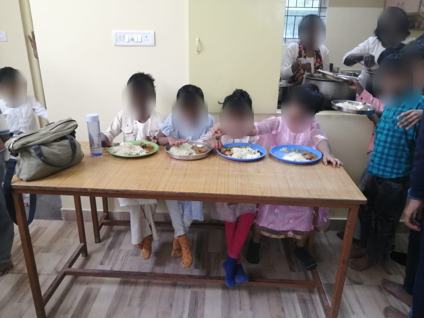 Four young children eating a meal at a table while staff serve from a hatch behind. Faces are obscured to protect the children's privacy.