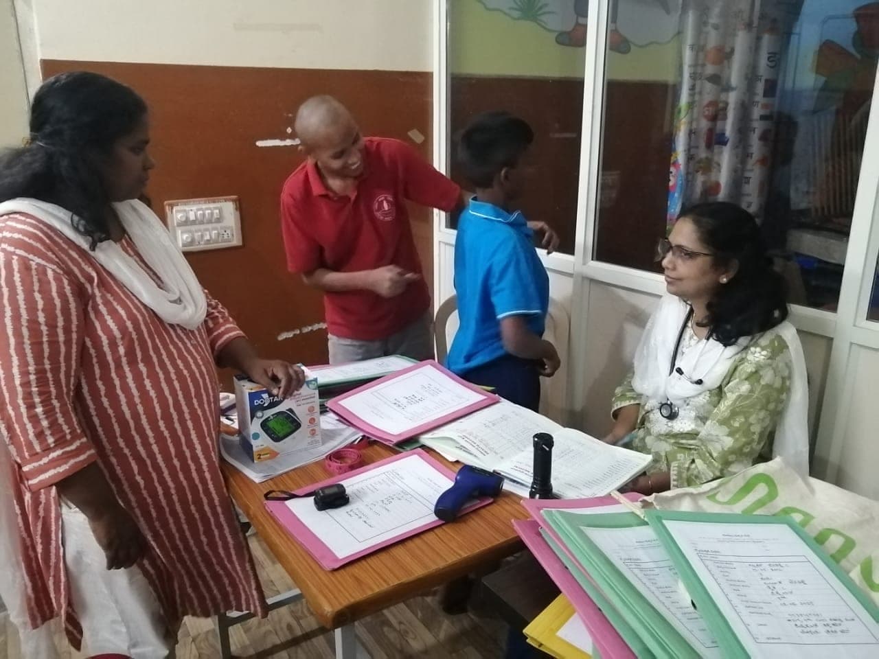 A doctor with a stethoscope seated at a desk of medical records while a child is measured.