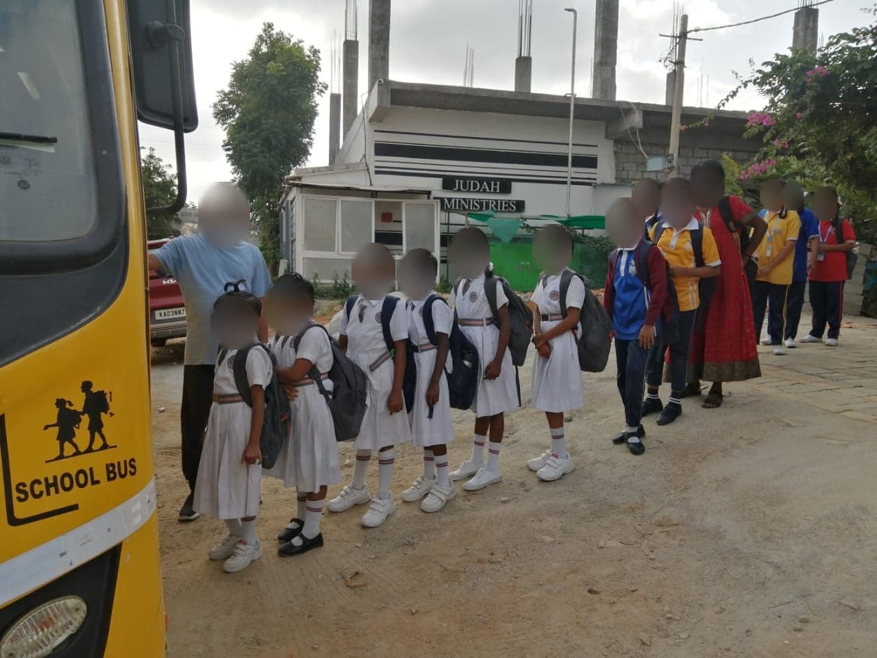 Children in white school uniform lined up beside a yellow school bus with an adult. Faces are obscured to protect the children's privacy.
