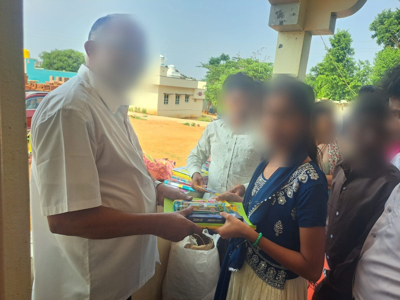 A student receiving a geometry set and notebooks, with others waiting behind. Faces are obscured to protect the children's privacy.