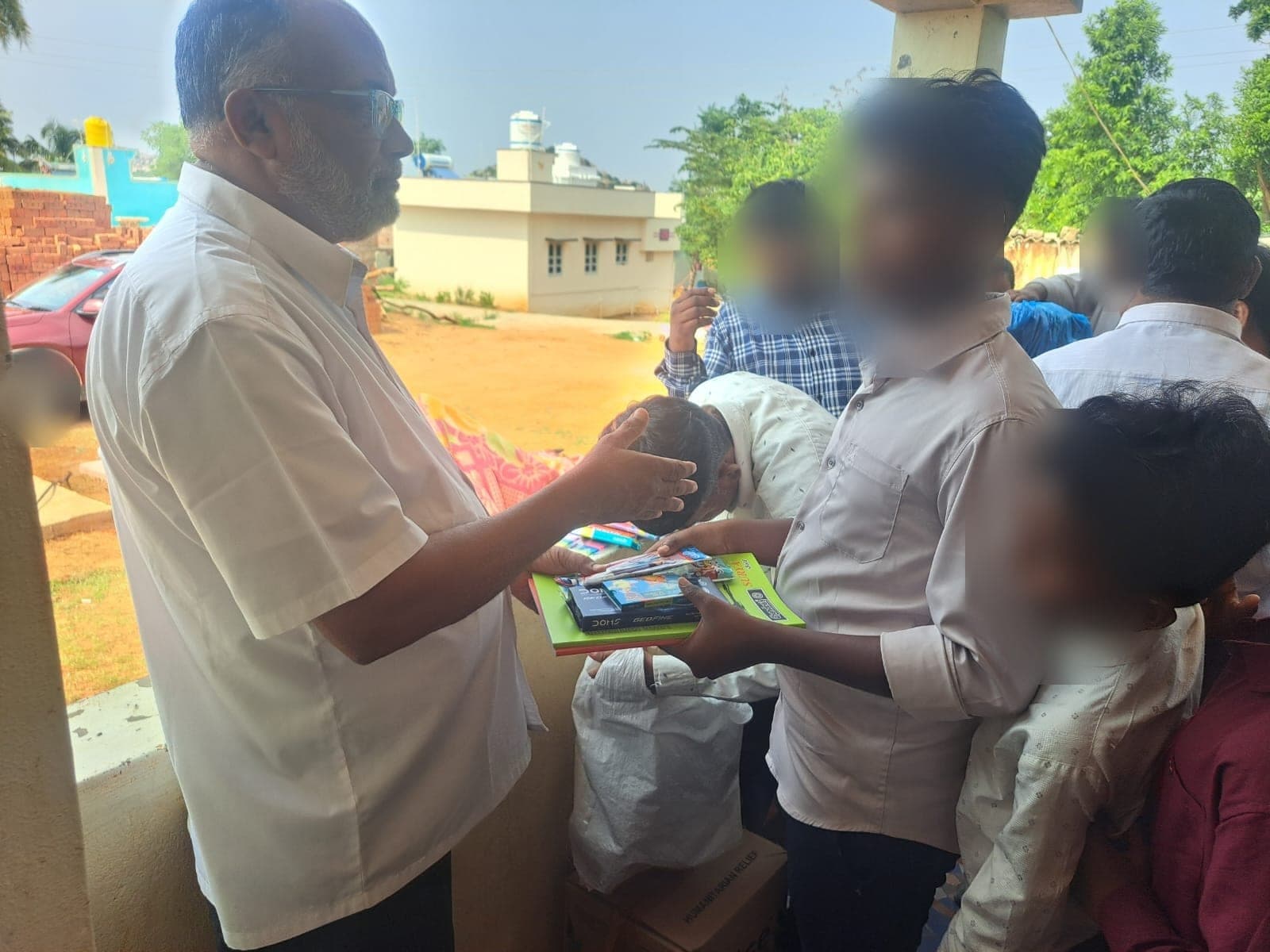 The founder resting a hand on a child's head in blessing while handing over school books. Faces are obscured to protect the children's privacy.