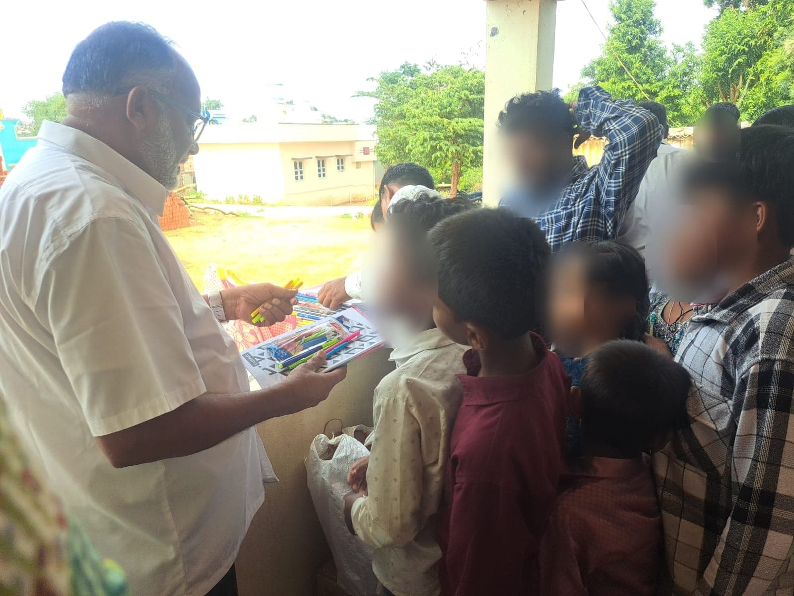 Children gathered closely around the founder as sets of colour pens are handed out. Faces are obscured to protect the children's privacy.