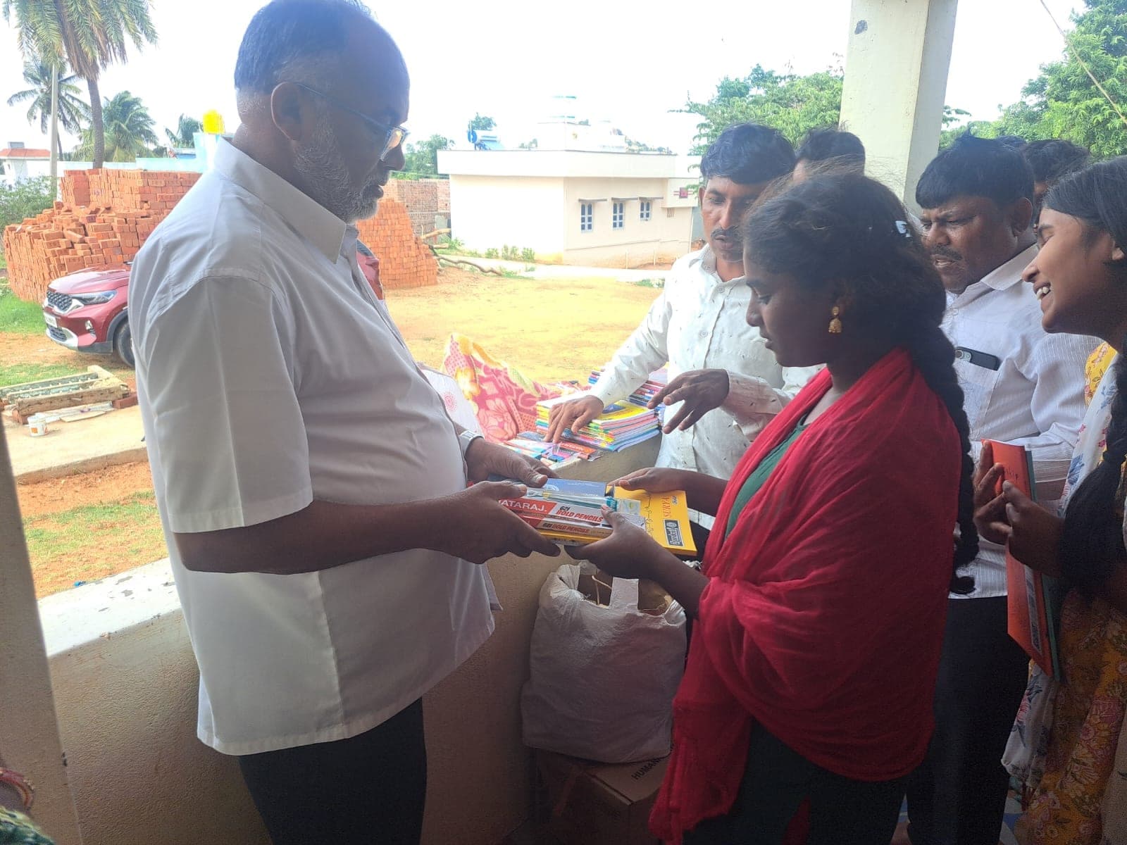The founder passing a set of pencils and notebooks to a student outside a village building.